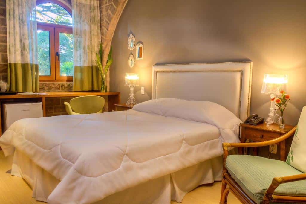 Pousada Castello Benvenutti no Vale dos Vinhedos com cama de casal bem ampla, cabeceira estofada, poltrona, mesa de trabalho com frigobar e abajures nas mesas de cabeceira