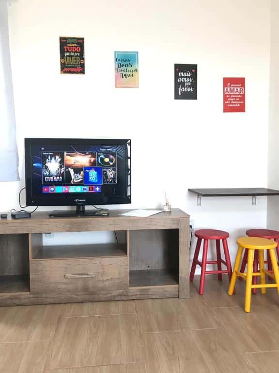 Uma TV em um suporte de madeira exibe opções de streaming; banquetas coloridas e arte na parede são vistas em uma sala bem iluminada e arrumada da Pousada Tchê Aprochega.