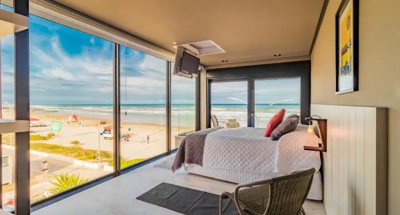 Quarto com janelas grandes com vista para a praia, com uma cama, cadeira e TV na parede.