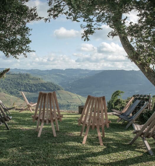 Foto de paisagem do Vale dos Vinhedos, no Rio Grande do Sul, com muita grama verde no chão, montanhas ao fundo, e cadeiras de madeira vazias no jardim, em primeiro plano