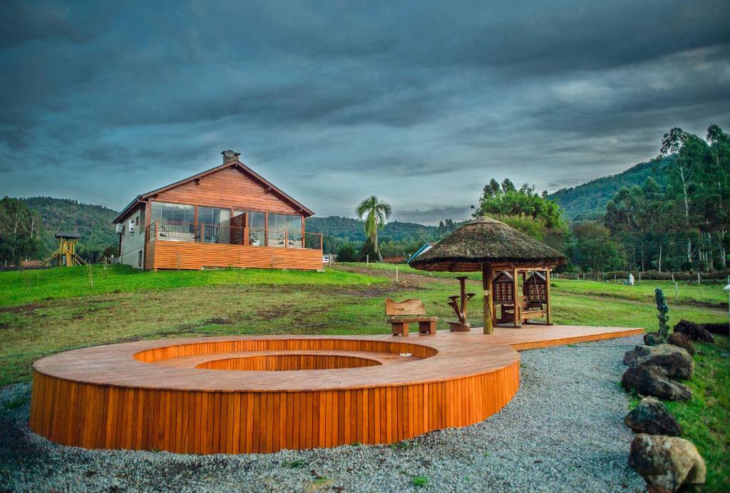 Uma casa de madeira com grandes janelas fica em uma colina gramada perto de um deck circular de madeira e de um gazebo com telhado de palha no Bangalôs Refúgio do Vale em Santa Maria do Herval.