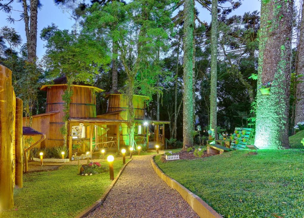 Um caminho de cascalho leva a uma casa de madeira rústica cercada por árvores e iluminada por luzes amarelas e verdes ao anoitecer, no Chalés Temático em Pipa de Vinho em Canela.