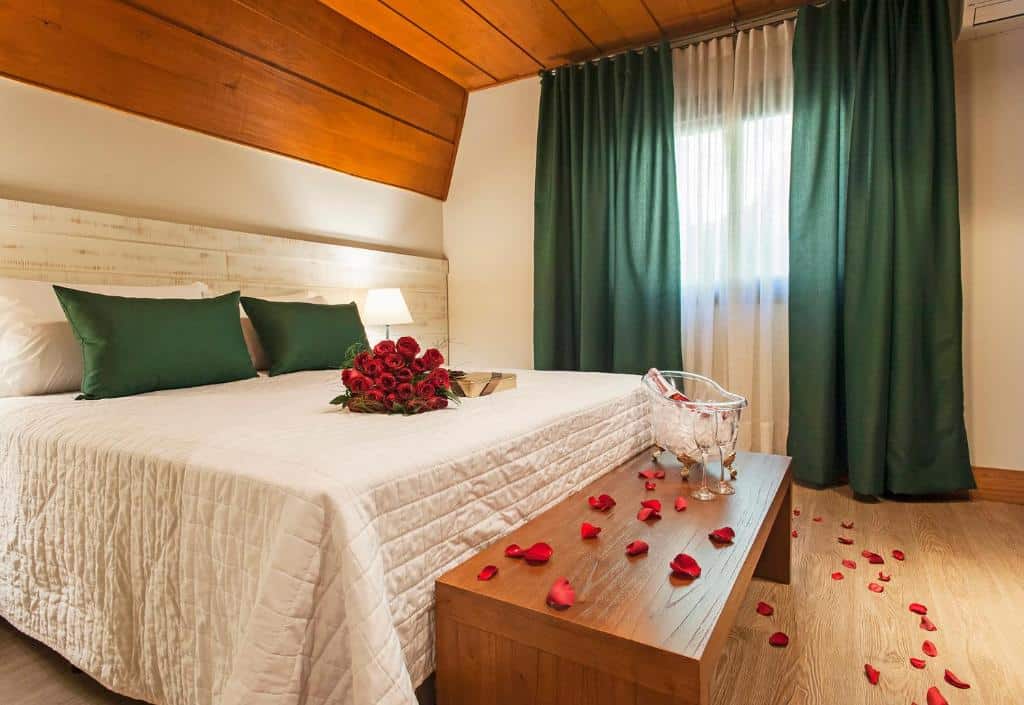 Uma cama com roupa de cama branca, travesseiros verdes, pétalas de rosas e um buquê de rosas vermelhas em um quarto com detalhes em madeira e cortinas verdes, nos Chalés Família Fioreze em Gramado.