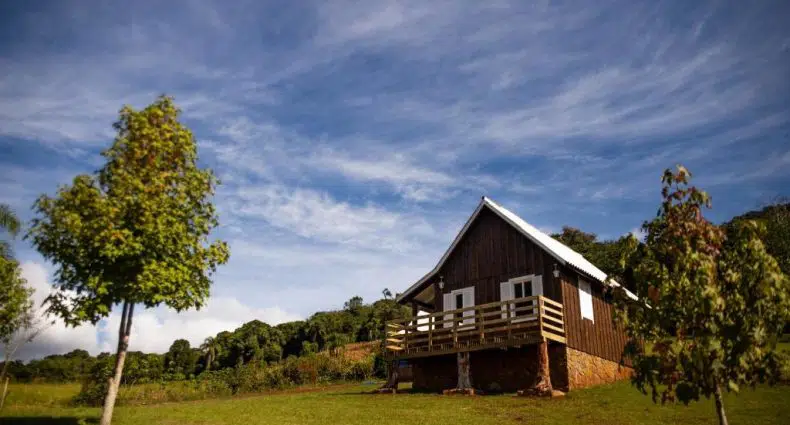 Uma pequena casa de madeira com uma varanda fica em uma colina gramada, com árvores e um céu parcialmente nublado ao fundo.