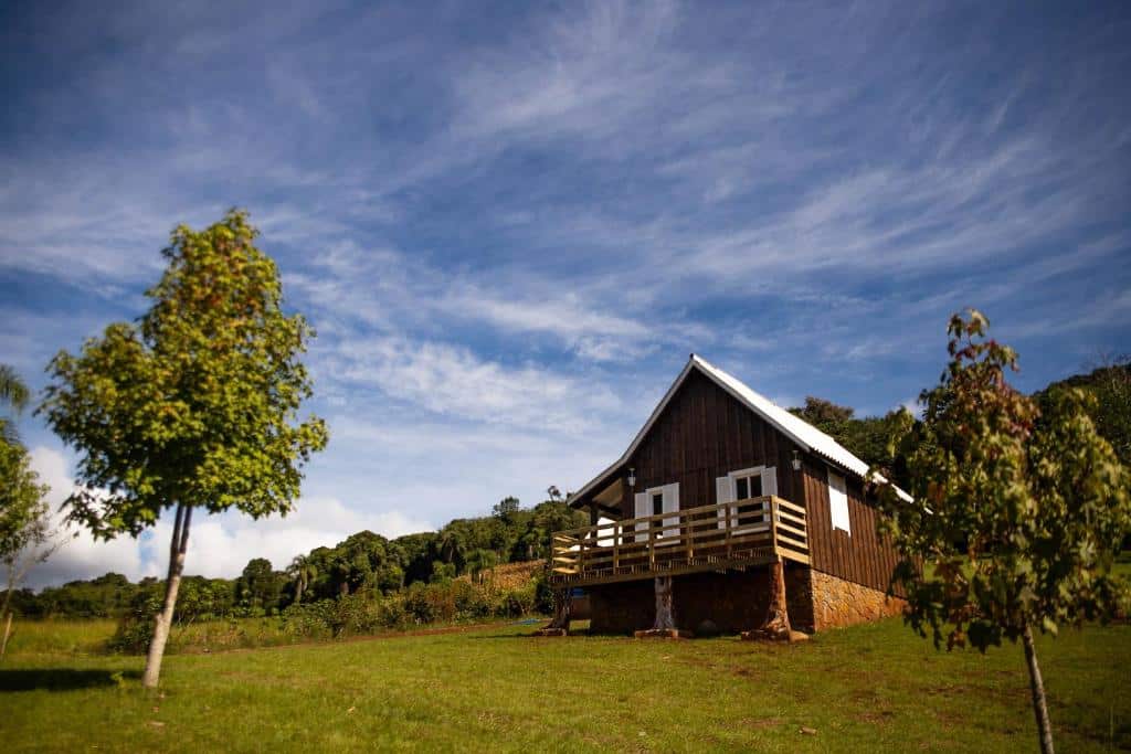 Uma pequena casa de madeira com uma varanda fica em uma colina gramada, com árvores e um céu parcialmente nublado ao fundo, nos chalés românticos no Rio Grande do Sul.