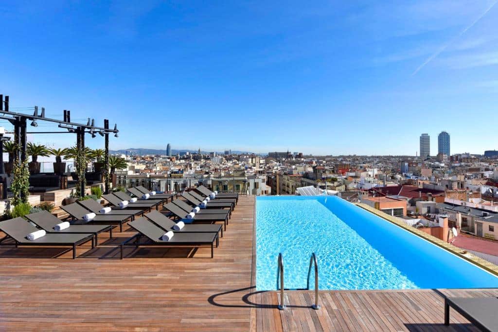 Piscina na cobertura do Grand Hotel Central, uma das recomendações de hotéis em Barcelona. Há várias espreguiçadeiras encarando a piscina, e a vista é para a cidade ao redor.