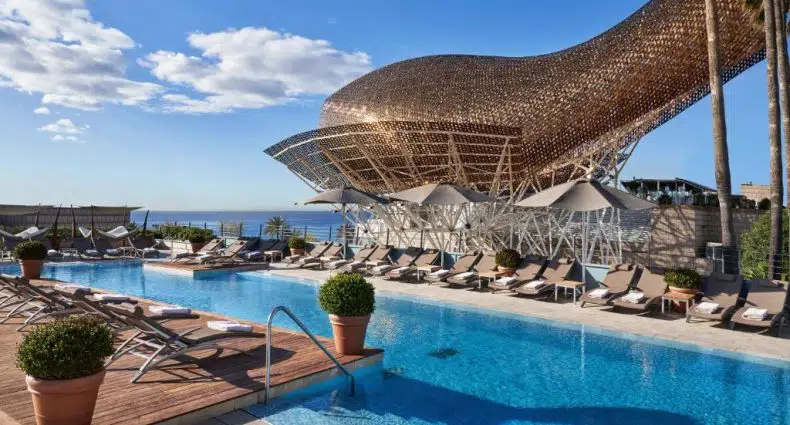 Piscina do Hotel Arts Barcelona, uma das recomendações de hotéis em Barcelona. Há uma escultura dourada atrás das espreguiçadeiras que encaram a piscina. Do outro lado, um deque de madeira tem mais espreguiçadeiras e algumas plantas.