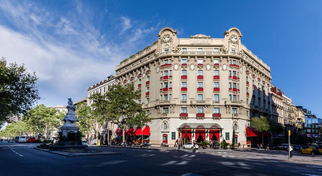 Fachada do El Palace Barcelona, um dos melhores hotéis em Barcelona. O hotel é bege com cortinas vermelhas na entrada. Há carros e pessoas circulando ao redor. O céu é azul acima.