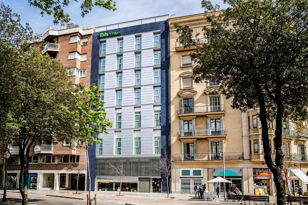 Fachada do ibis Styles Barcelona Centre, uma das recomendações de hotéis em Barcelona. O prédio de seis andares tem o nome do hotel na parte de baixo e no topo. Há outros prédios ao lado, assim como algumas pessoas caminhando e árvores dos dois lados da foto.