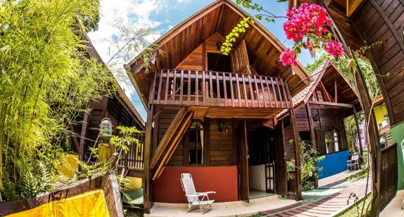 Uma cabana de madeira de dois andares com varanda, cercada por vegetação e flores, em um dia ensolarado.