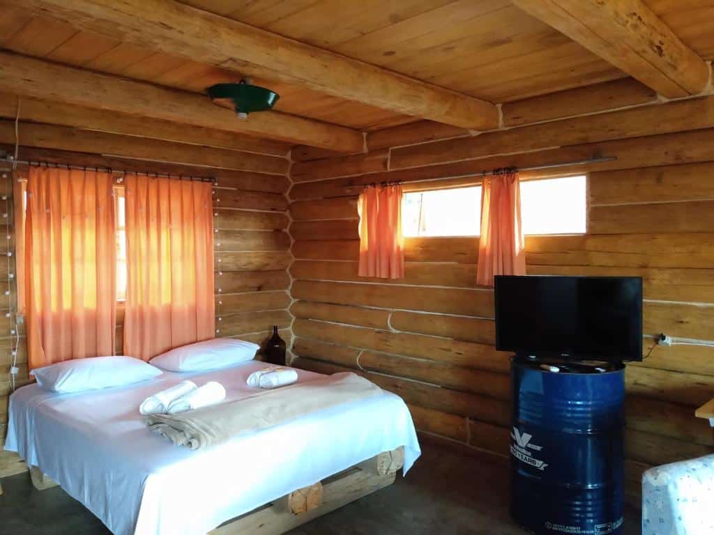 Um quarto de cabine de madeira com uma cama, cortinas cor de pêssego, uma TV em um barril azul e toalhas dobradas sobre a cama, no Morro do Vento em Picada Café, para representar chalés românticos no Rio Grande do Sul.