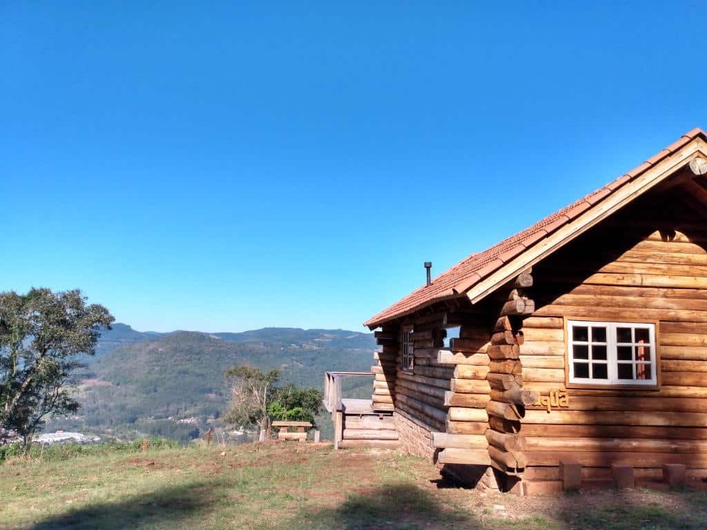 Uma cabana de madeira fica no topo de uma colina gramada com montanhas e céu azul claro ao fundo, no Morro do Vento em Picada Café.