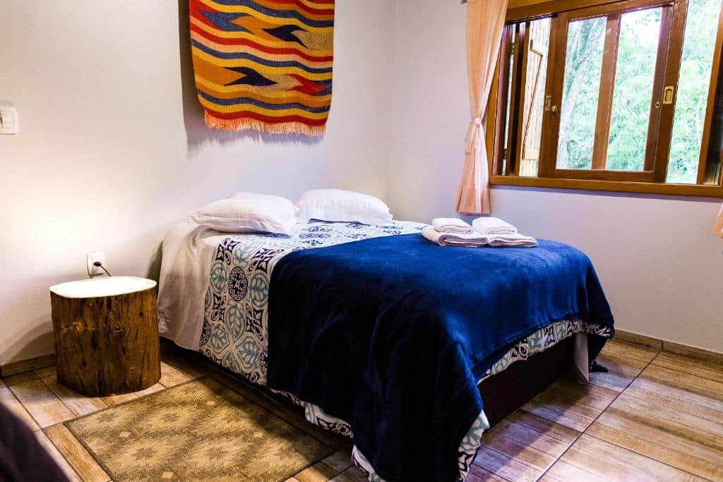 Uma cama bem arrumada no Ninho das Águias Cabana em Nova Petrópolis com roupa de cama azul, dois travesseiros e toalhas dobradas ao lado de uma janela com cortinas bege, para representar chalés românticos no Rio Grande do Sul.