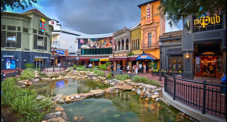 Área de compras e restaurantes ao ar livre com edifícios coloridos, um lago rochoso em primeiro plano e um céu nublado acima.