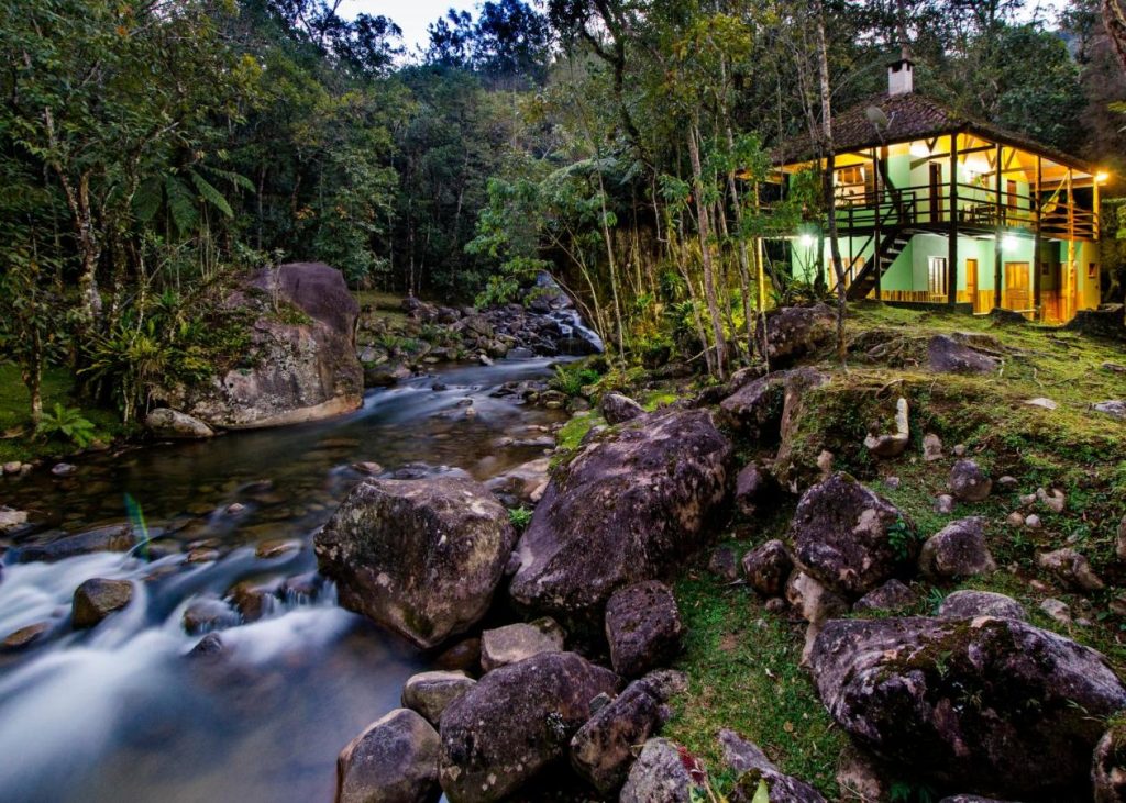 Riacho em movimento com muitas pedras, árvores verdes e uma casa de dois andares em tom de verde durante o dia, ilustrando post pousadas em Visconde de Mauá.