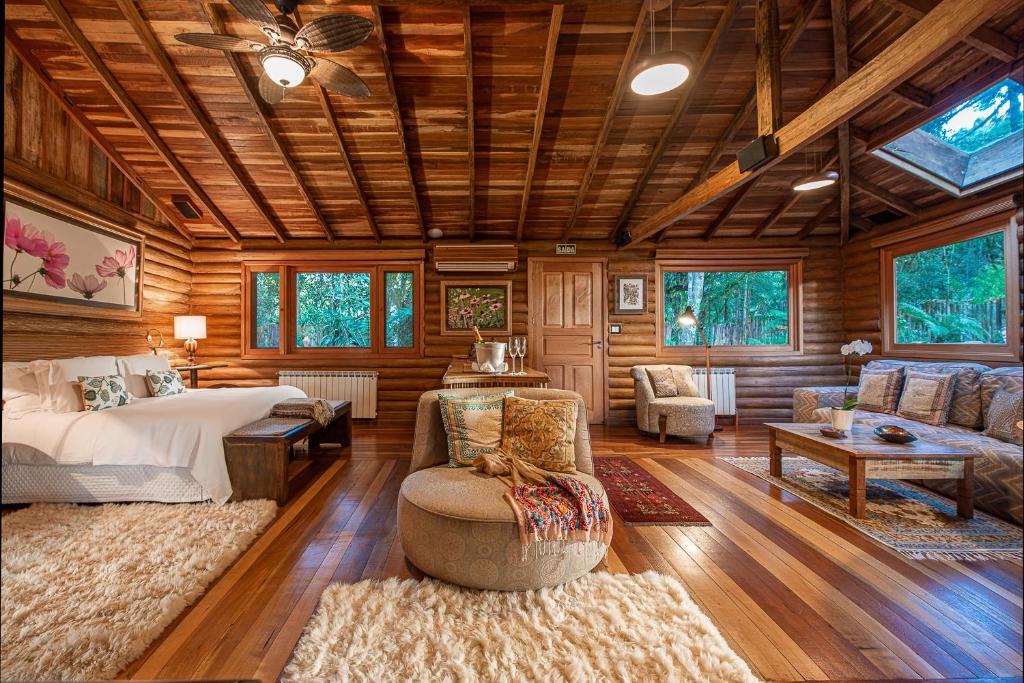 Um quarto espaçoso na Pousada do Engenho em São Francisco de Paula em uma cabana de madeira com cama, sofá, tapetes, teto de madeira e janelas grandes com vista para as árvores, para representar chalés românticos no Rio Grande do Sul.