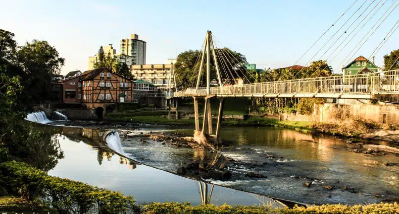 Uma ponte de pedestres suspensa por cabos cruza um rio com pequenas cachoeiras e edifícios ao fundo em um dia ensolarado, em Timbó, para representar pousadas no Valeu Europeu em Santa Catarina.