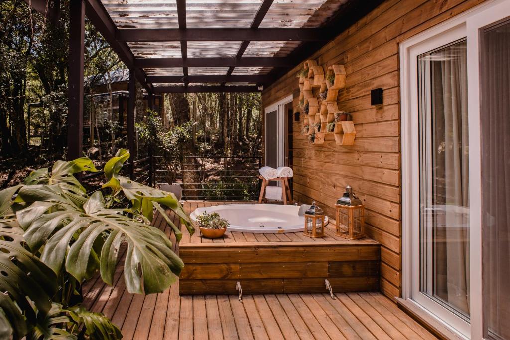 Deck de madeira com banheira de hidromassagem, vasos de plantas, lanternas e decoração de parede, anexado a uma casa com painéis de madeira em uma área de floresta, na Vila Nambu em Gramado.