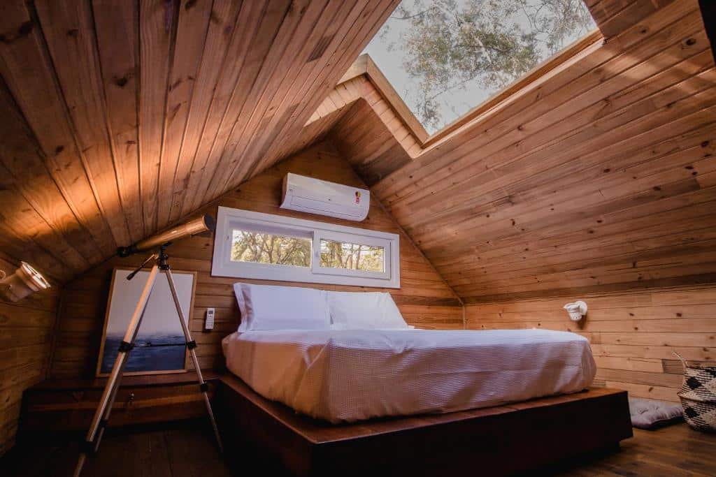 Um quarto aconchegante na Vila Nambu em Gramado, no sótão com painéis de madeira, uma cama com lençóis brancos, uma claraboia e um telescópio ao lado da janela, para representar chalés românticos no Rio Grande do Sul.