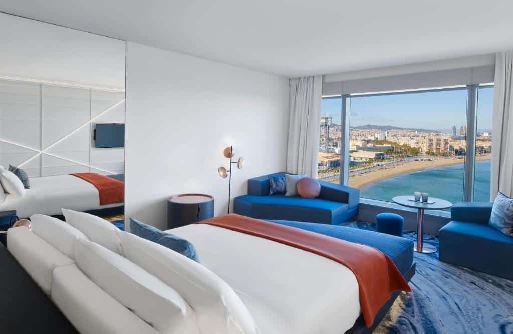 Quarto do W Barcelona. A cama de casal encara uma janela que tem vista para a praia La Barceloneta. Há dois sofás do lado da janela, e uma mesinha no centro. Um espelho está na parede ao lado da cama.