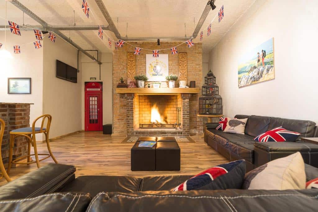 lounge compartilhado do Britânico Upstairs com dois sofás de couro escuro, almofadas com o símbolo da inglaterra e uma grande lareira de tijolos no centro