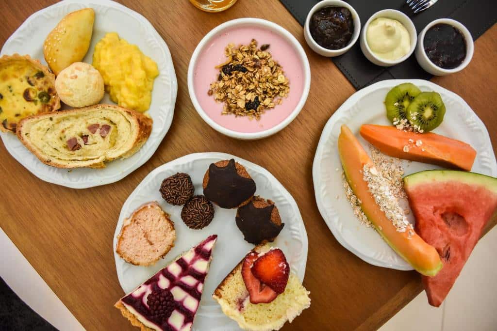 mesa de café da manhã do Micheline Hotel Tricot com três pratos repletos de alimentos (frutas, pães doces e salgados, tortas doces e salgadas), uma tigela de iogurte e três potinhos de molhos ao lado.
