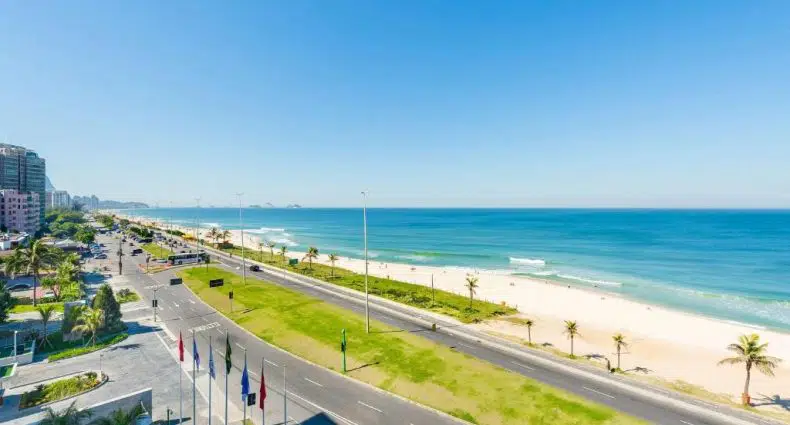 Uma estrada larga corre paralela a uma praia de areia e ao oceano azul sob um céu claro, com prédios e palmeiras nas proximidades.