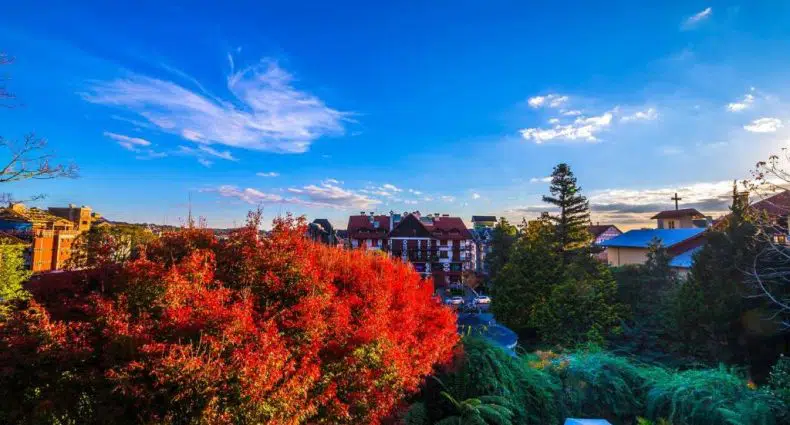 Folhagem vermelha de outono em primeiro plano, telhados e edifícios no centro, céu azul com nuvens dispersas acima. Essa é o Hotel Casa da Montanha, um dos hotéis na serra gaúcha.
