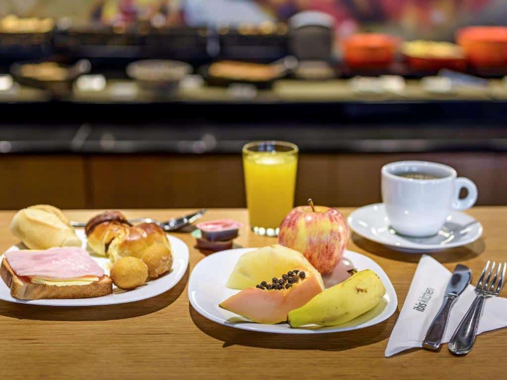 Pratos do café da manhã no ibis Guarulhos, com frutas, pães e frios, além de um copo de suco de laranja e uma xícara com café, sendo que há talheres posicionados ao lado direito