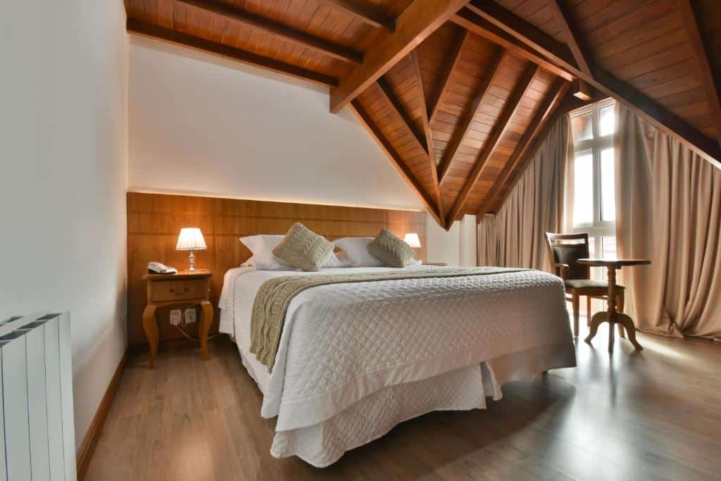 quarto do Micheline Hotel Tricot com uma cama de casal ao centro, uma pequena mesa de madeira redonda com uma cadeira em frente às janelas amplas e teto de madeira aparente.