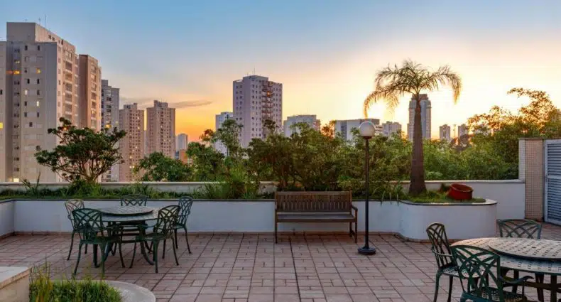 Pátio externo com mesas e cadeiras de metal, um banco, plantas e prédios da cidade visíveis ao pôr do sol ao fundo.