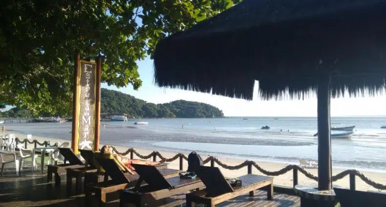 Espreguiçadeiras de madeira sob um telhado de palha estão voltadas para a praia de areia e o mar calmo, com barcos e árvores ao fundo na Pousada Estrela do Mar.
