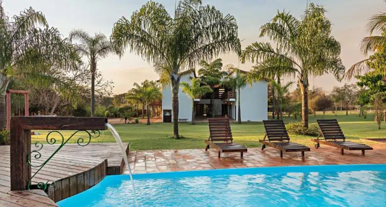 Uma piscina com espreguiçadeiras e palmeiras em frente a uma casa branca moderna em um gramado.