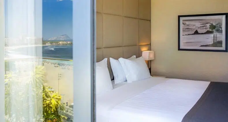 Uma cama bem arrumada com roupas de cama brancas ao lado de uma janela com vista para o litoral; arte emoldurada pendurada na parede.