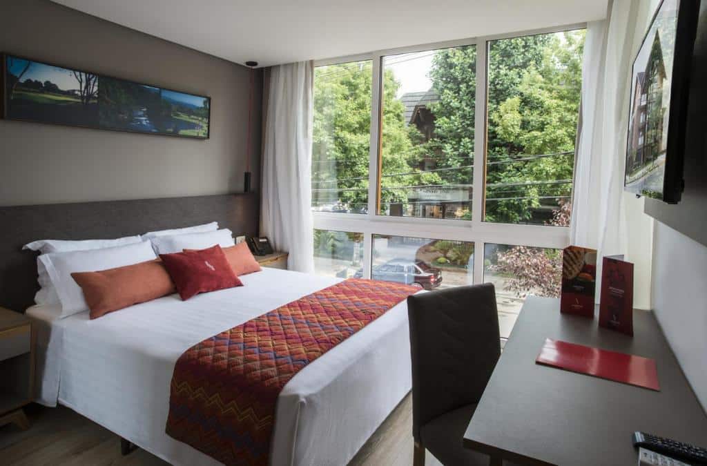 quarto do Hotel Laghetto Stilo Centro em Gramado com uma cama de casal no lado esquerdo e uma mesa em frente. Ao fundo há uma parede completamente coberta por janelas de vidro que dão visão para a rua