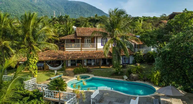 Uma casa de dois andares com telhado de telha, cercada por palmeiras, tem uma piscina e um deck de madeira em primeiro plano, no O Vila Toro Guest House, para representar pousadas de luxo em São Paulo.