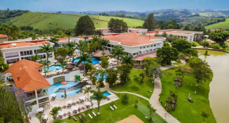 Vista do grande Vale Suíço Resort com telhados vermelhos, piscinas, palmeiras e jardins, cercado por colinas e um lago sob um céu claro. Representa uma das opções de resorts em Minas Gerais.