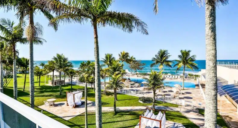 Palmeiras e espreguiçadeiras cercam uma área de piscina com vista para a praia e o oceano sob um céu azul claro.