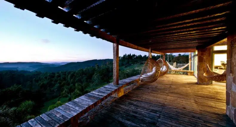 Um deck de madeira com cadeiras de rede tem vista para uma paisagem montanhosa verde ao pôr do sol, parcialmente coberto por um telhado de madeira.