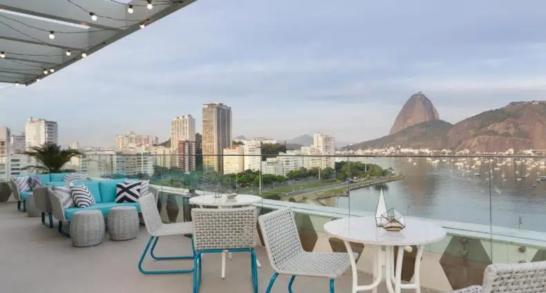 O terraço externo com mesas e cadeiras brancas tem vista para uma baía, prédios da cidade e uma montanha sob um céu nublado, na propriedade do Yoo2 Rio de Janeiro by Intercity, para representar hotéis em Botafogo.