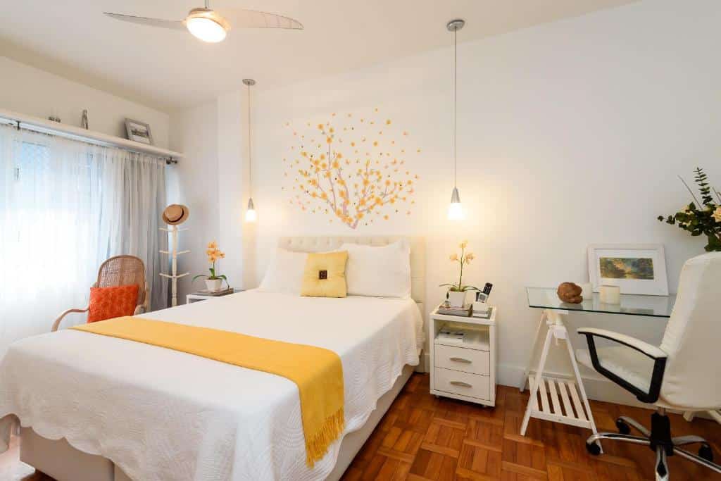 Copacabana Beach Amazing Apartment em Copacabana com quarto elegante com paredes brancas, desenho de ipê amarelo sobre a cabeceira, cama de casal com almofada e peseira amarelas, poltrona e cabideiro ao fundo, além de mesinhas de cabeceira e mesa de trabalho