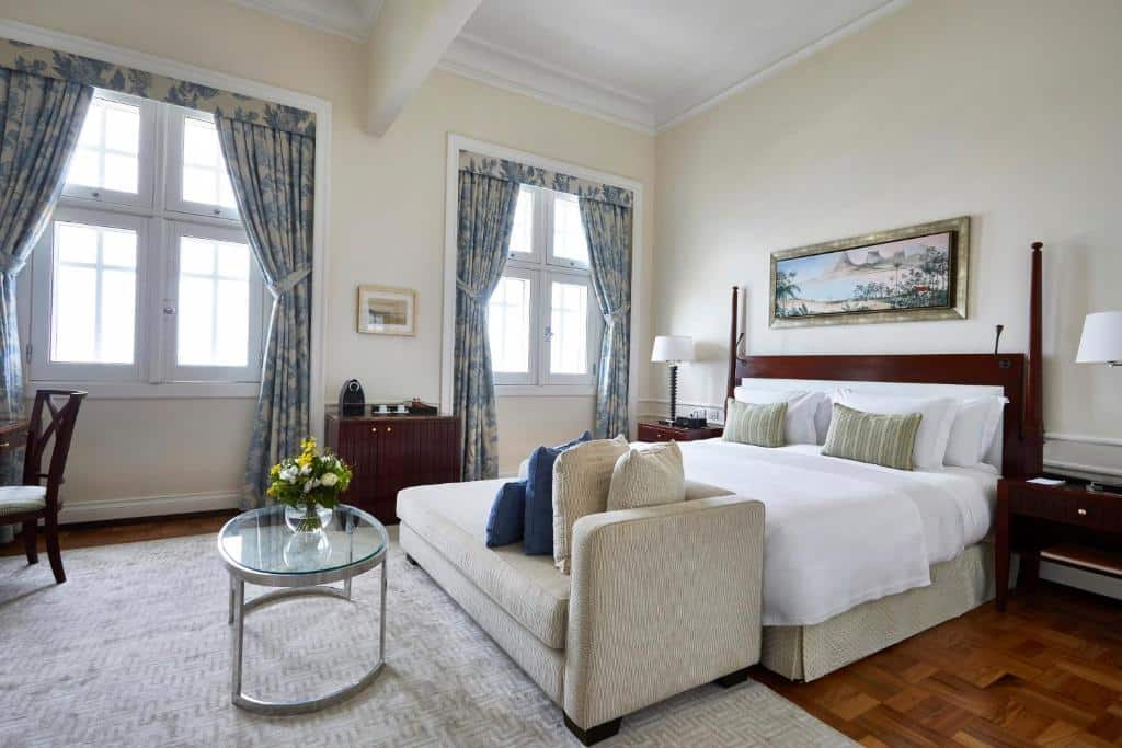 suíte do Copacabana Palace no rio de janeiro com cama e casal extragrande, quadro decorativo sobre a cabeceira, duas janelas com cortinas abertas, e um sofá aos pés da cama