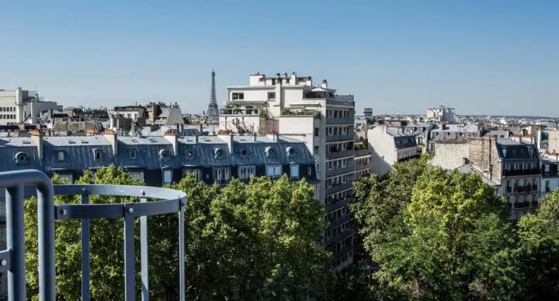 Vista da Torre Eiffel a partir do Mercure Paris 17 Batignolles e edifícios de Paris com árvores em primeiro plano e a Torre Eiffel visível à distância em um céu claro, para representa hotéis Mercure em Paris.