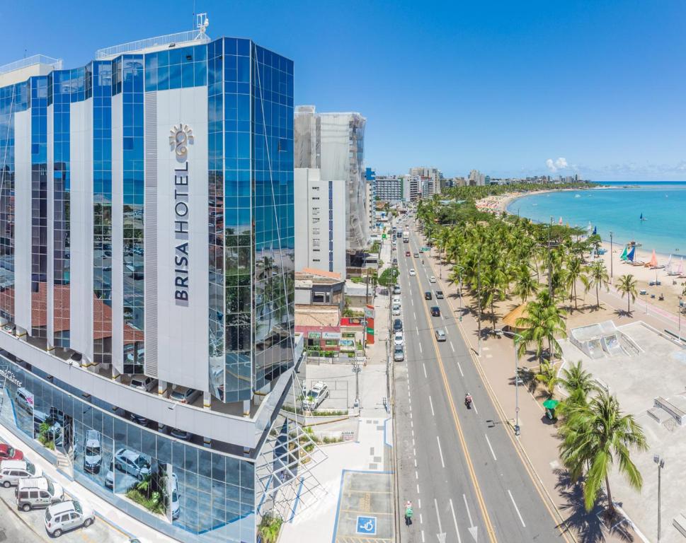 Vista do prédio de um dos hotéis em Maceio, sendo o edificil espelhado escrito na vertical "Brisa Hotel" com o desenho de um sol ao lado. O local fica ao lado de uma avenida movimentada com carros andando e estacionados, e com a praia ao lado com mar azul e coqueiros na orla