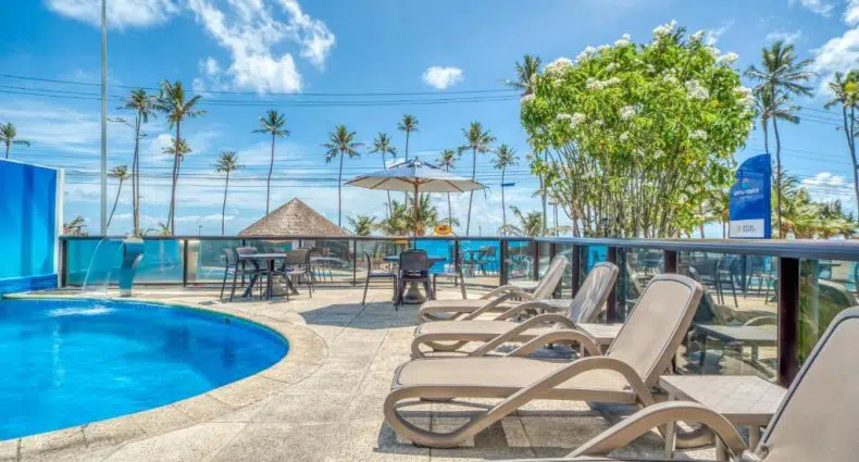 Espreguiçadeiras em uma piscina externa curva com mesas, guarda-sóis, palmeiras e um céu azul ao fundo.