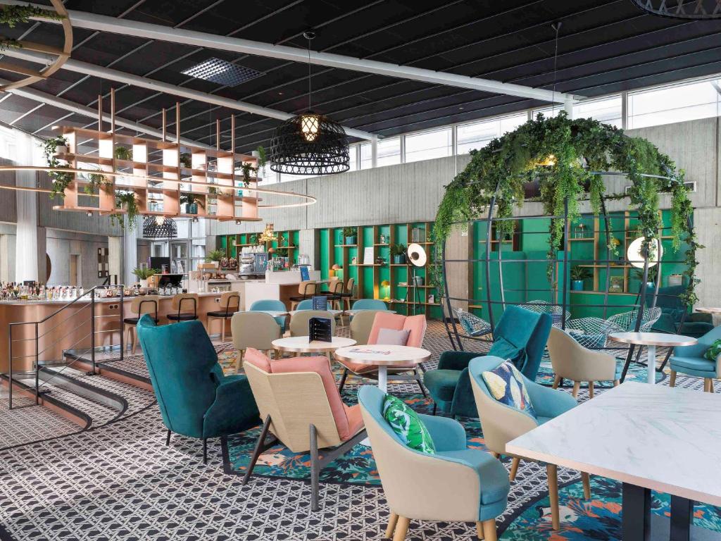 Restaurante do Novotel Paris Charles de Gaulle Airport decorado em tons pastéis com mesas e cadeiras estofadas espalhadas. Ao fundo fica o bar.