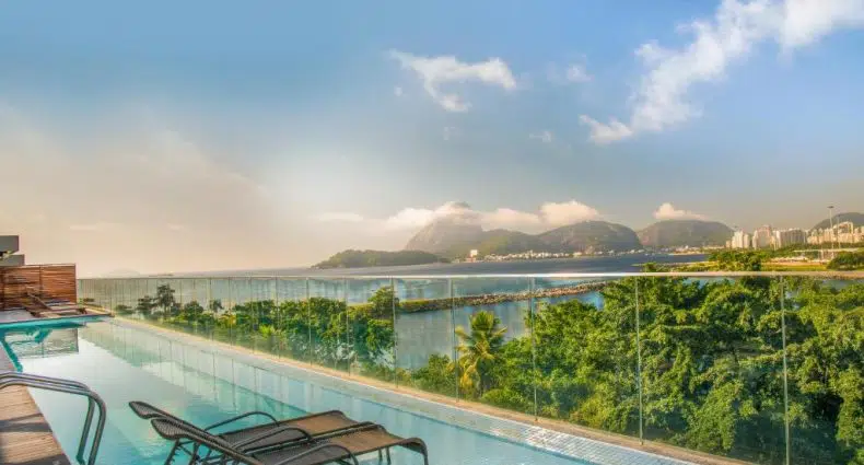 Piscina infinita na cobertura com espreguiçadeiras, guarda-corpo de vidro e vista para a vegetação, água e montanhas distantes sob um céu azul.
