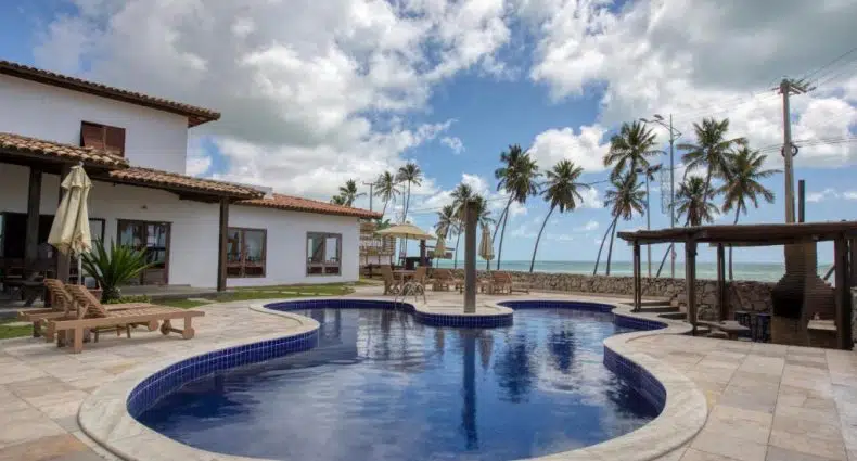 Piscina externa com bordas curvas cercada por espreguiçadeiras, palmeiras e um prédio próximo sob um céu parcialmente nublado, na propriedade da Pousada Coqueiral, para representar pousadas em Maceió.