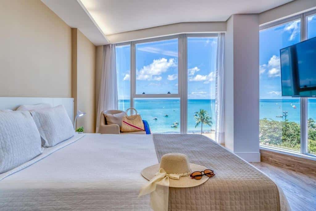 quarto do Hotel Brisa Suites, com uma cama de casal com um chapéu de praia e óculos em cima, uma Tv, uma poltrona com uma bolsa de palha em cima e, ao fundo, tem paredes de vidro com vista panorâmica do mar