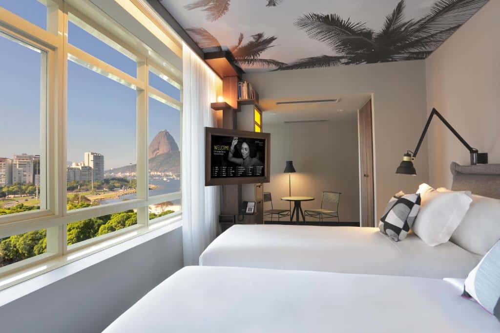 quarto do yoo2 com duas camas de solteiro, teto decorado com palmeiras, TV presa à parede perto das camas, mesa de trabalho ao fundo, e vista para o RJ da ampla janela em frente às camas